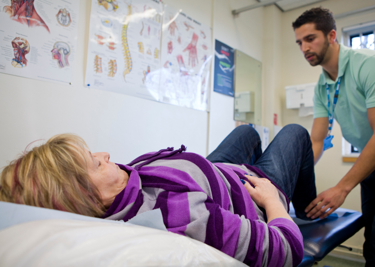 Male short haired Physiotherapist Holds The Ankles Of A  female Patient who lays on a therapy bed with their knees bent. 