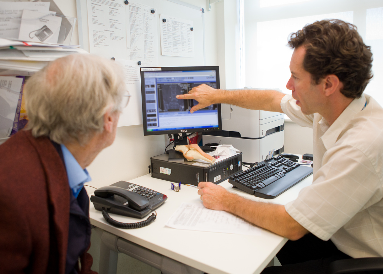Clinician Sitting At Desk Next To An Elderly Patient And Pointing At His Computer Screen which features and xray of his knee.