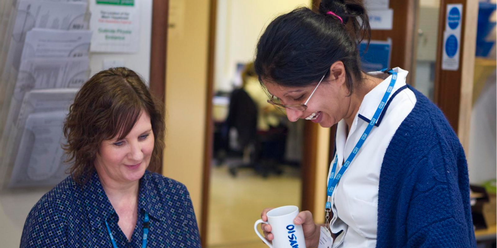 A member of staff smiling and sitting at a desk with another member of staff standing next to her and laughing whilst holding a mug. 